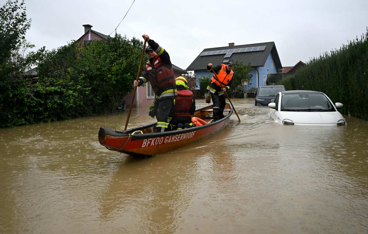 Men in high-vis paddling a small red boat through floodwaters, two cars submerged. 
