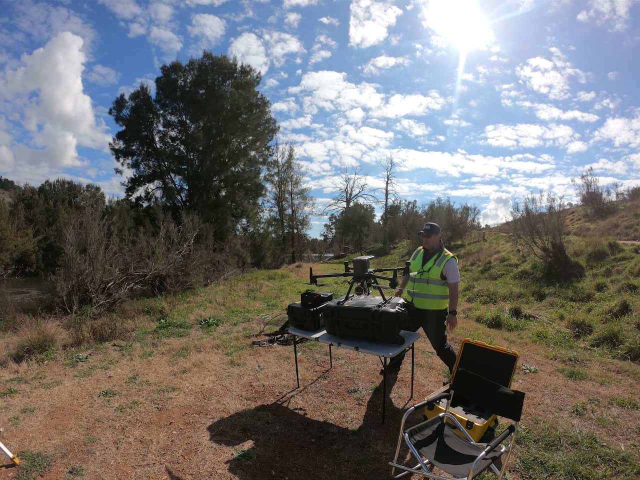 A man wearing a hi-vis vest in a small clearing in the bush, stands next to a small fold-up table with a large drone on it.