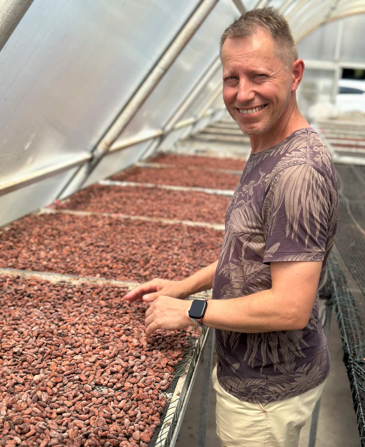 A man in a brown T-shirt stands in front of trays filled with cacao beans.