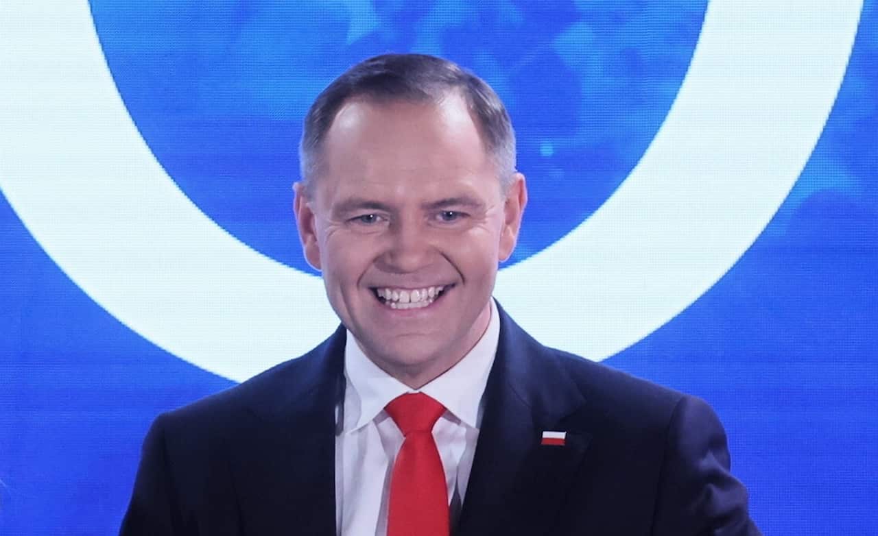 A man in a suit with a Polish flag pin on it, standing in front of a blue background.