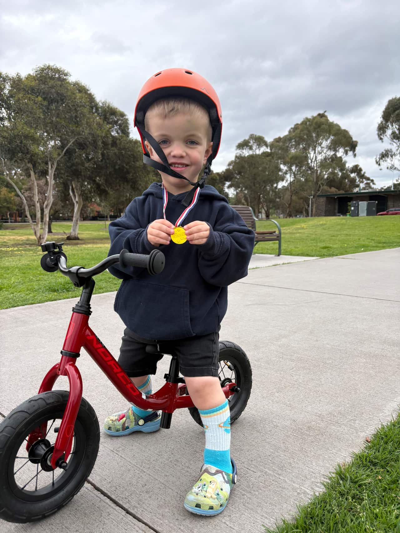 A young smiling boy sits on a bicycle with no pedals. He is wearing an helmet and holding a gold medal, which is hung around his neck. 