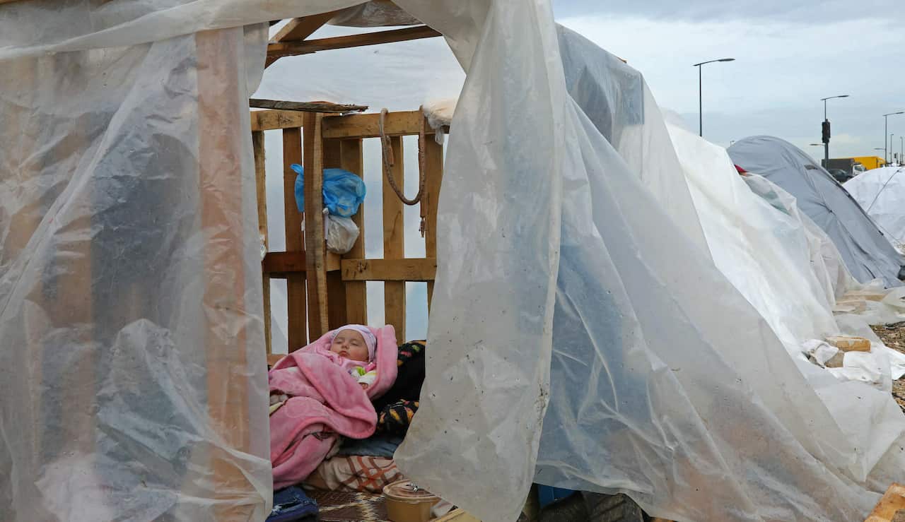 A baby wrapped in a pink blanket lies on a makeshift bed inside a tent made of wooden pallets and transparent plastic sheets under a cloudy sky.