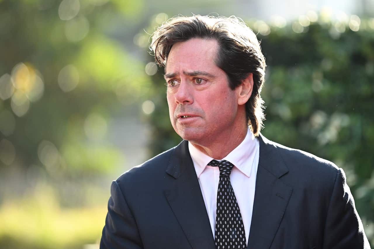 Man standing in front of greenery wears grey suit and black tie. 