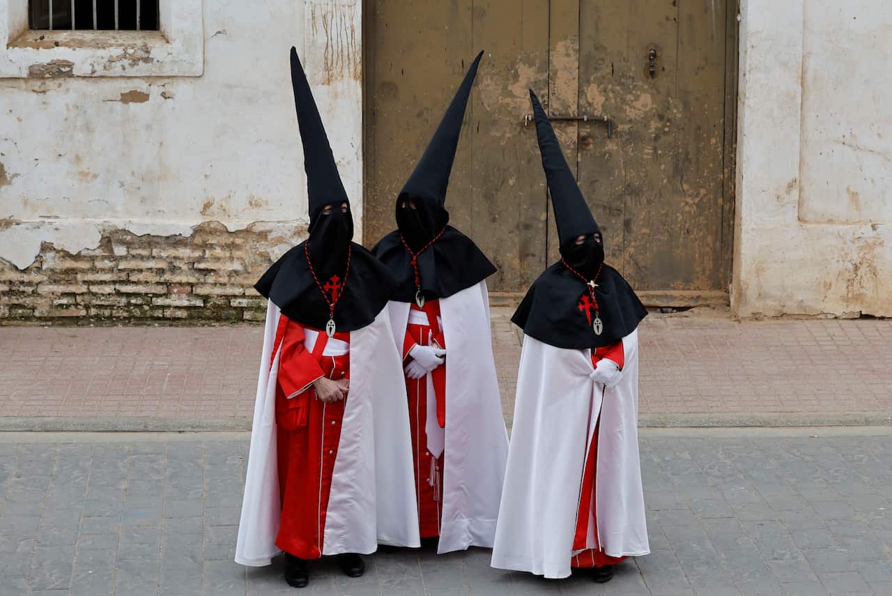 Several people wearing white and red robes with tall black pointy hoods stand in front of a water-damaged building.