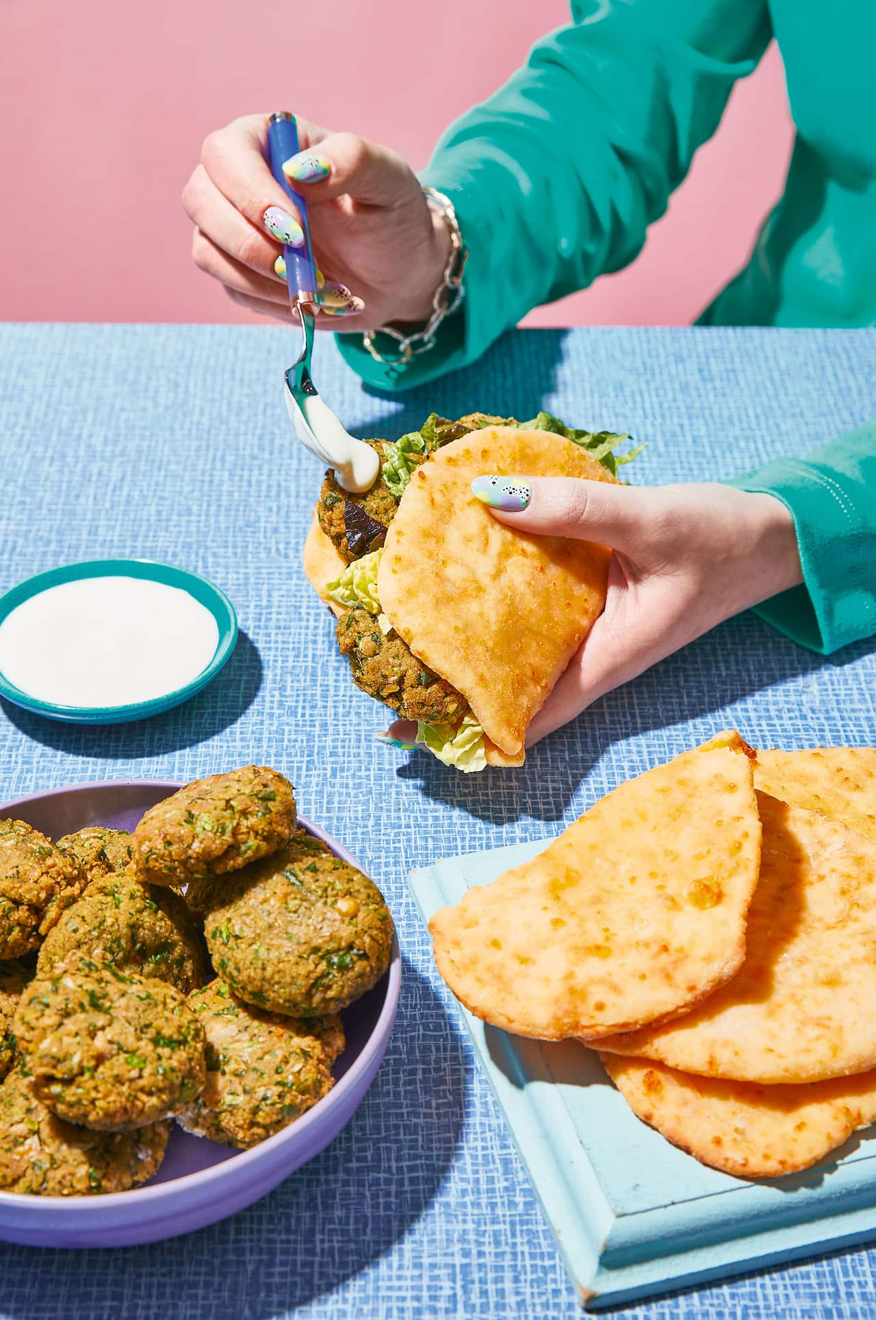 A hand holds a flatbread folded around a filling. A bowl of falafel and a board with more flatbreads sits on the table. 