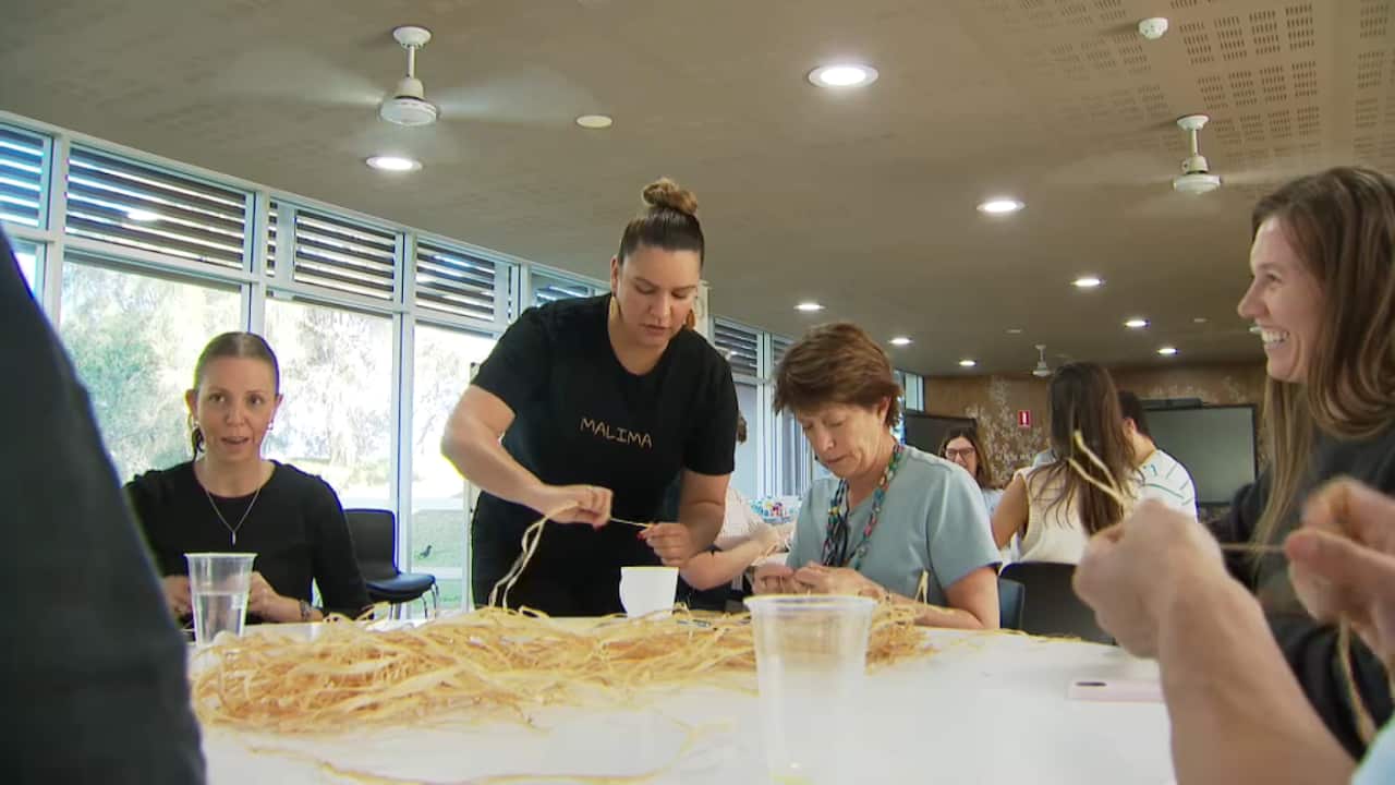 A group gathers at a weaving workshop facilitated by Malima at La Perouse in coastal Sydney. Attendees are laughing, smiling and staring down in concentration while weaving.