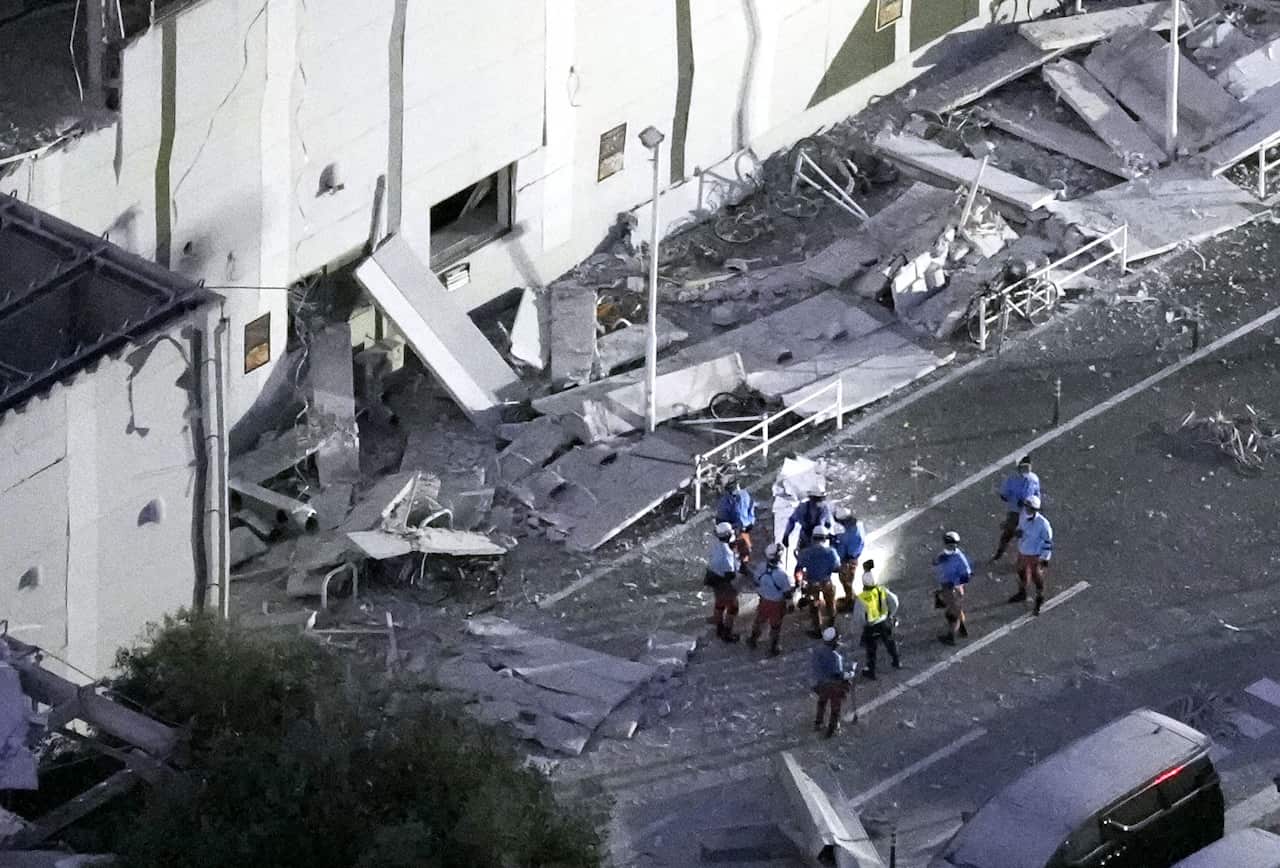A group of people in blue vests and hard hats stand in front of a building. An awning close to the building has collapsed, leaving debris strewn across the ground.