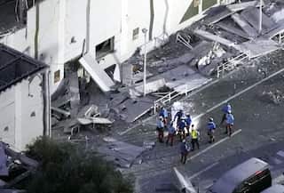 A group of people in blue vests and hard hats stand in front of a building. An awning close to the building has collapsed, leaving debris strewn across the ground.