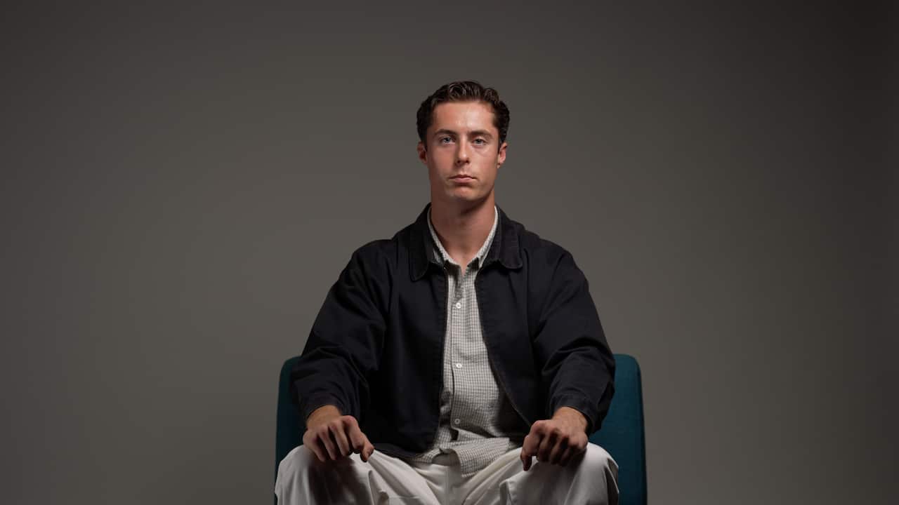 a young man wearing a button down shirt and a black jacket sits on a couch with his hands on his lap with a grey background