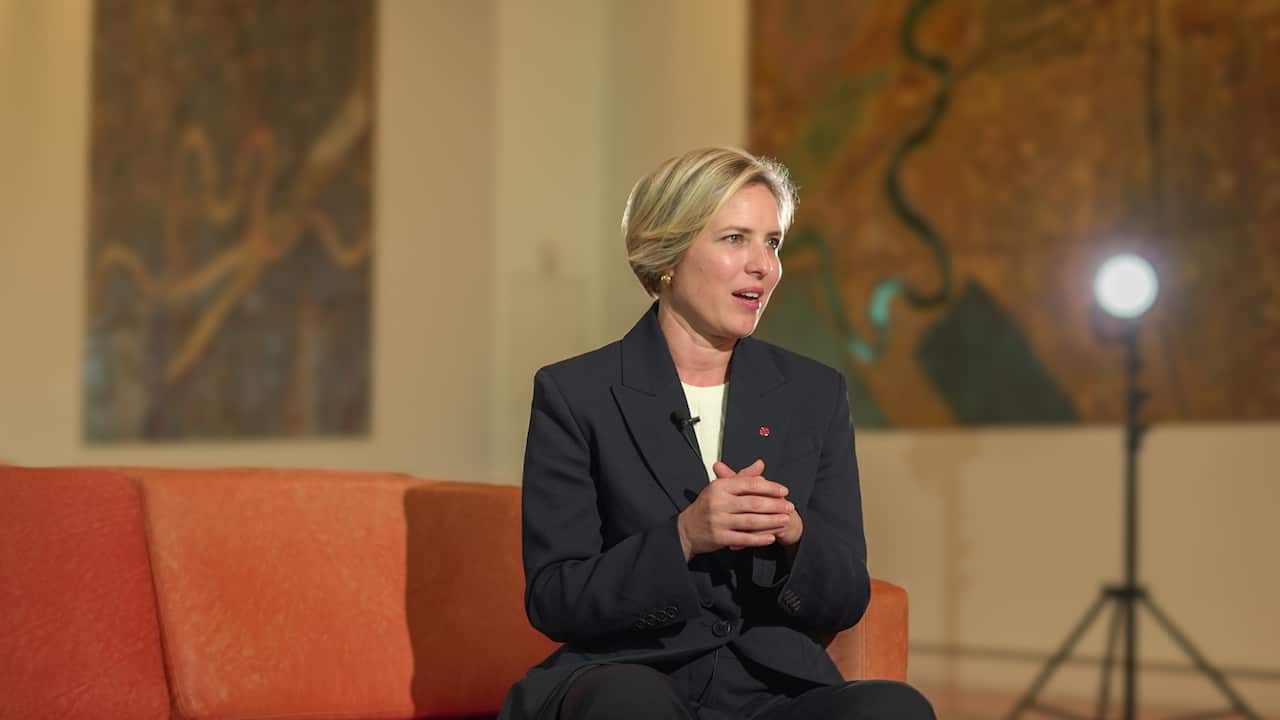 A blonde woman in a navy suit, sitting on an orange couch.
