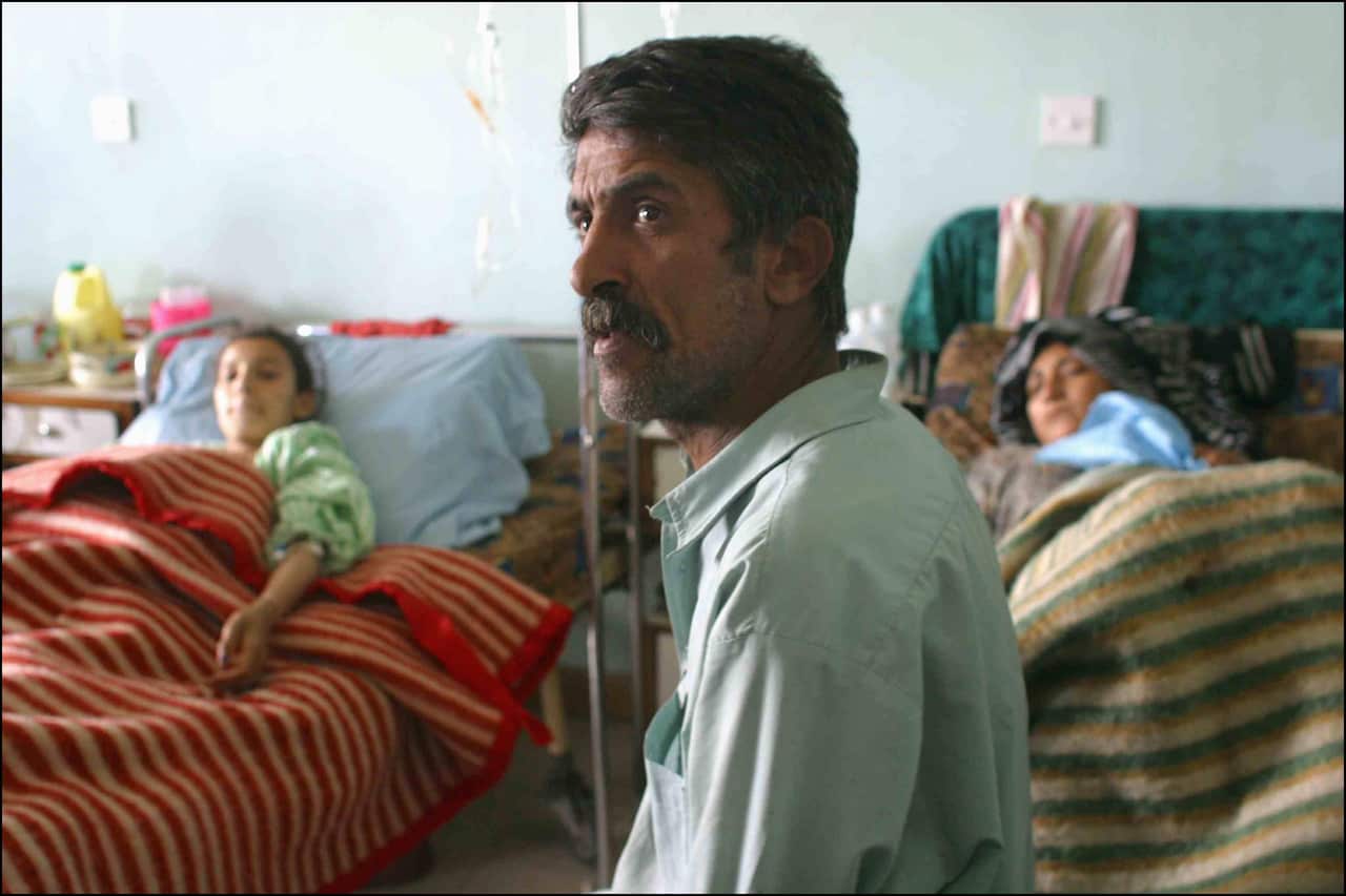 A man sits in a hospital alongside two people in beds.
