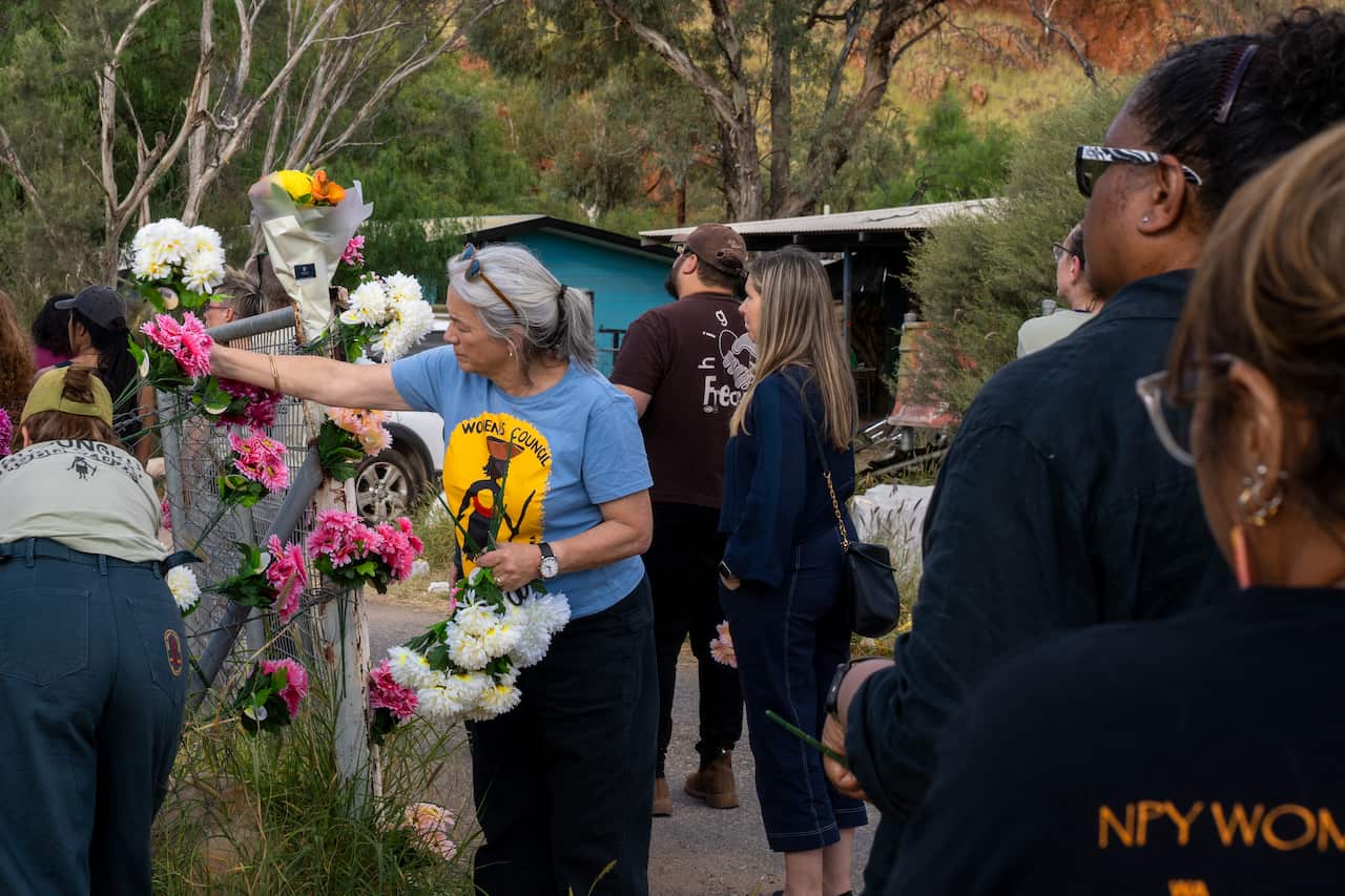 A woman places flowers into a chain link fence in a rural landscape with tin-roofed houses in the background.