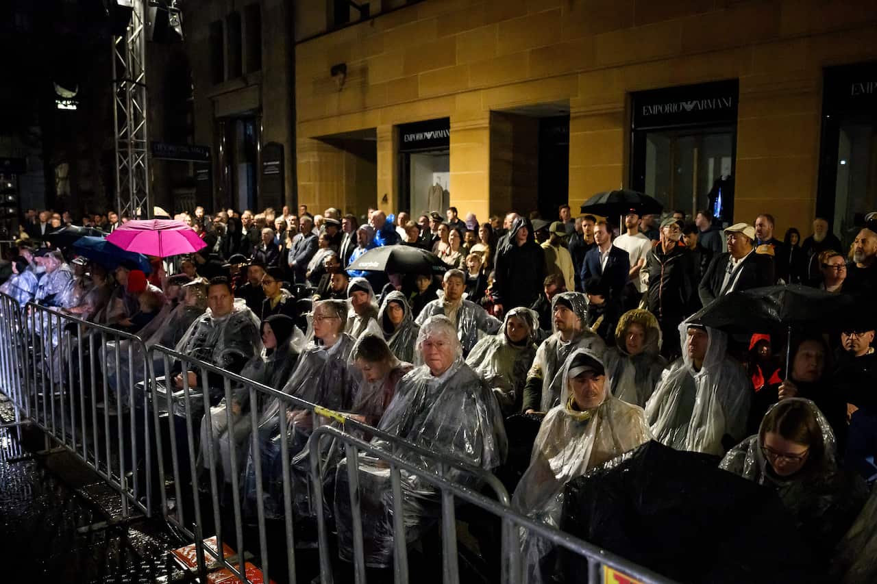 A large crowd of people, sitting down behind a railing, many in rain ponchos.