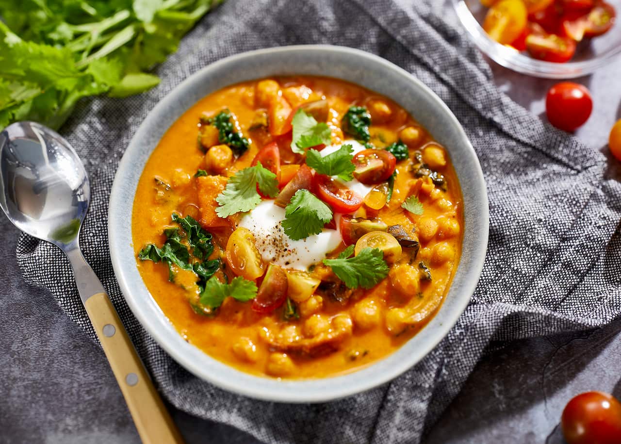 A pale bowl sits on a grey-toned cloth napkin. It holds a vibrant orange-yellow soupy stew, studded with chickpeas and vegetables. A metal soup spoon with a cream handle sits alongside.