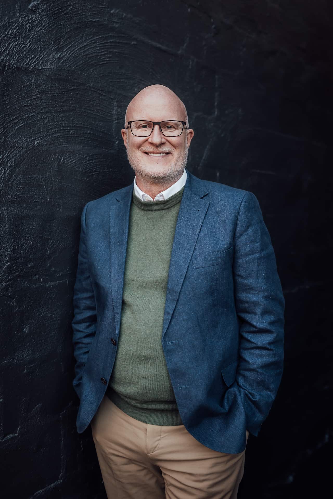 A man in a blue blazer worn over a green jumper is smiling.