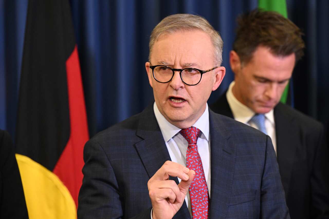 Prime Minister Anthony Albanese wearing a suit and tie and talking inside. There is an Aboriginal flag behind him.