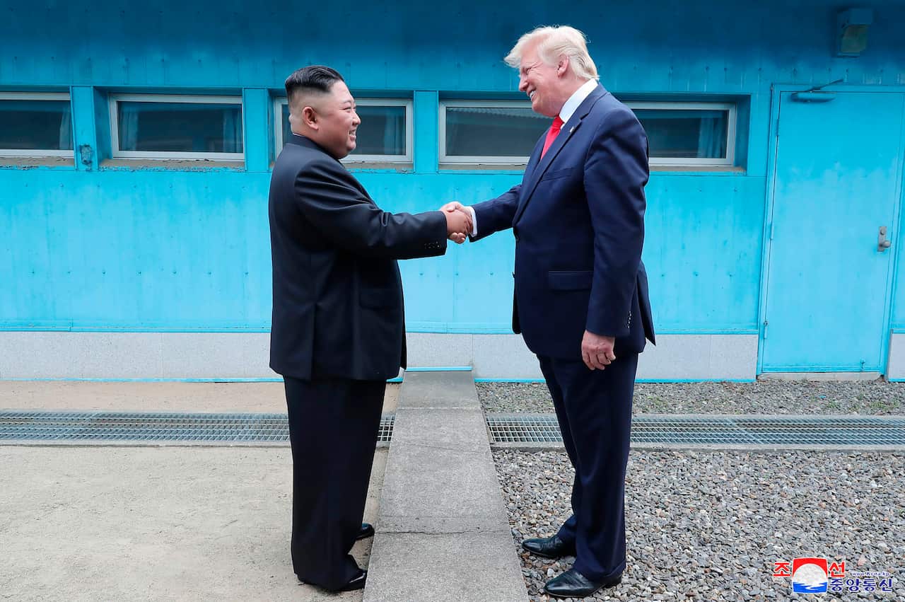 Kim Jong Un and Donald Trump shake hands over a concrete line in front of a blue building.