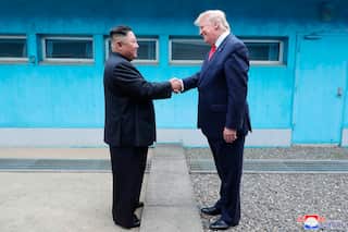 Kim Jong Un and Donald Trump shake hands over a concrete line in front of a blue building.