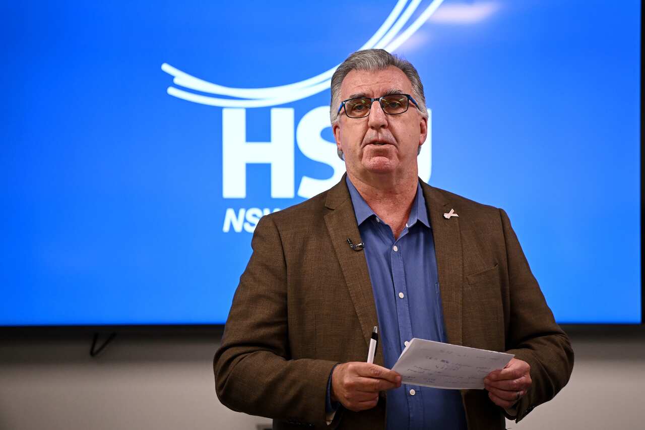 A man in a suit and wearing glasses. Behind him is a large blue screen with HSU NSW written on it in white lettering. 