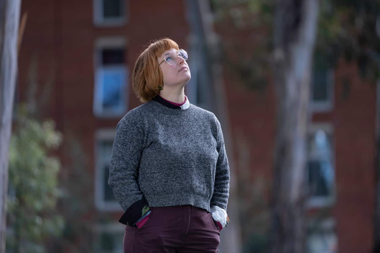 A woman in a blue jumper and glasses looks up at the sky with a blurry building behind her.