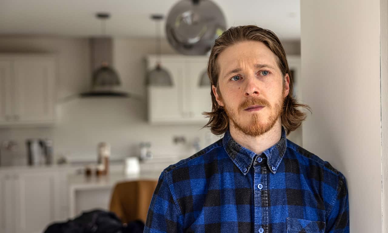 A man in a blue checked shirt stands in a kitchen.