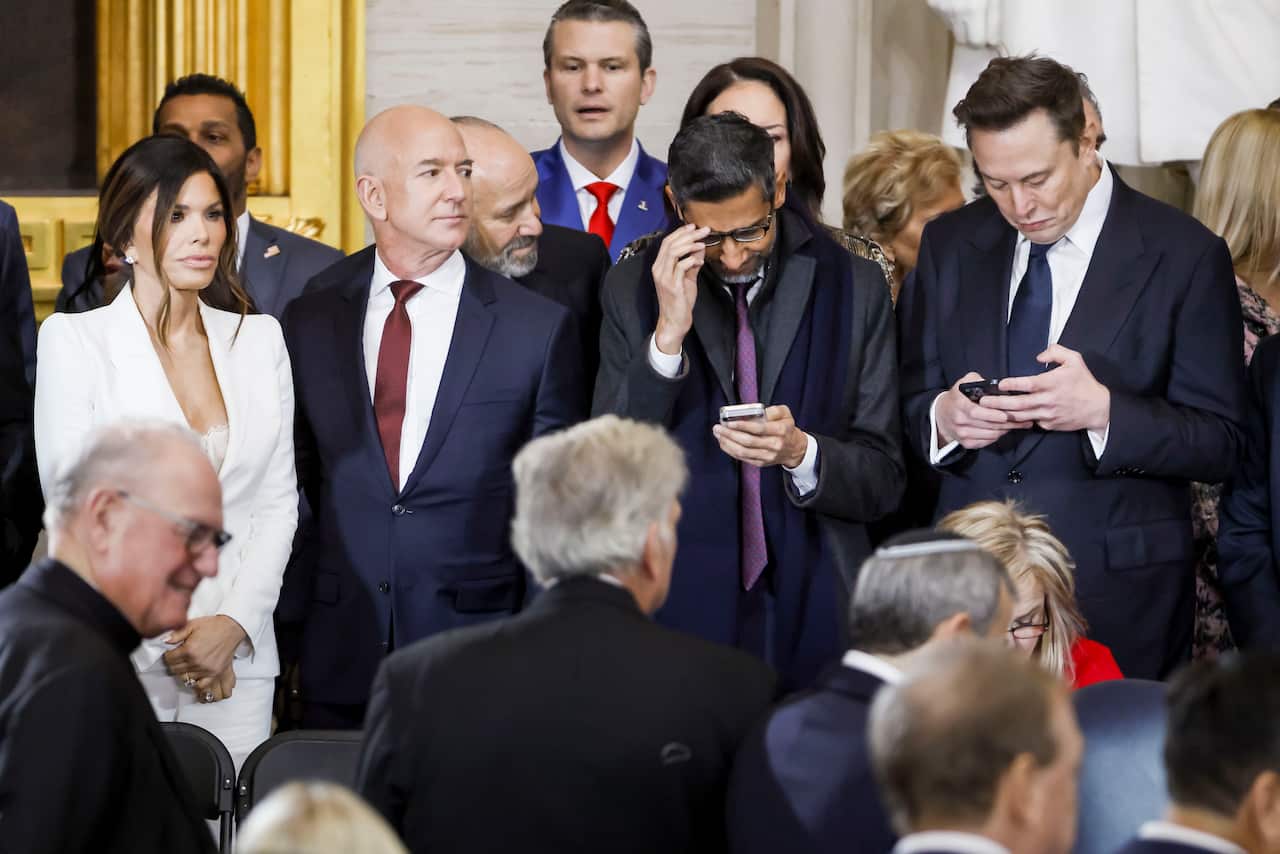 A crowd dressed in formal attire stands, with some looking at their phone screens.
