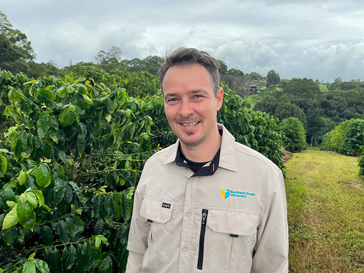 A man in a khaki shirt stands in a field next to green trees. 