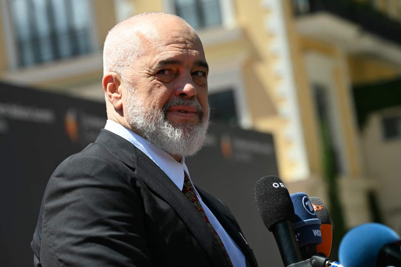 A man with a white beard and wearing a suit speaks in front of microphones outdoors in Montenegro.