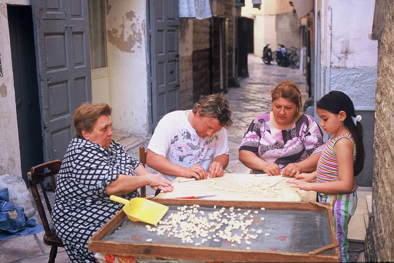 Jamie Oliver sits at a table in a laneway with two women and a young girl, making pasta. 