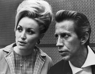 Black-and-white portrait of two people seated side by side, looking off-camera, with distinctive voluminous hairstyles and patterned clothing.
