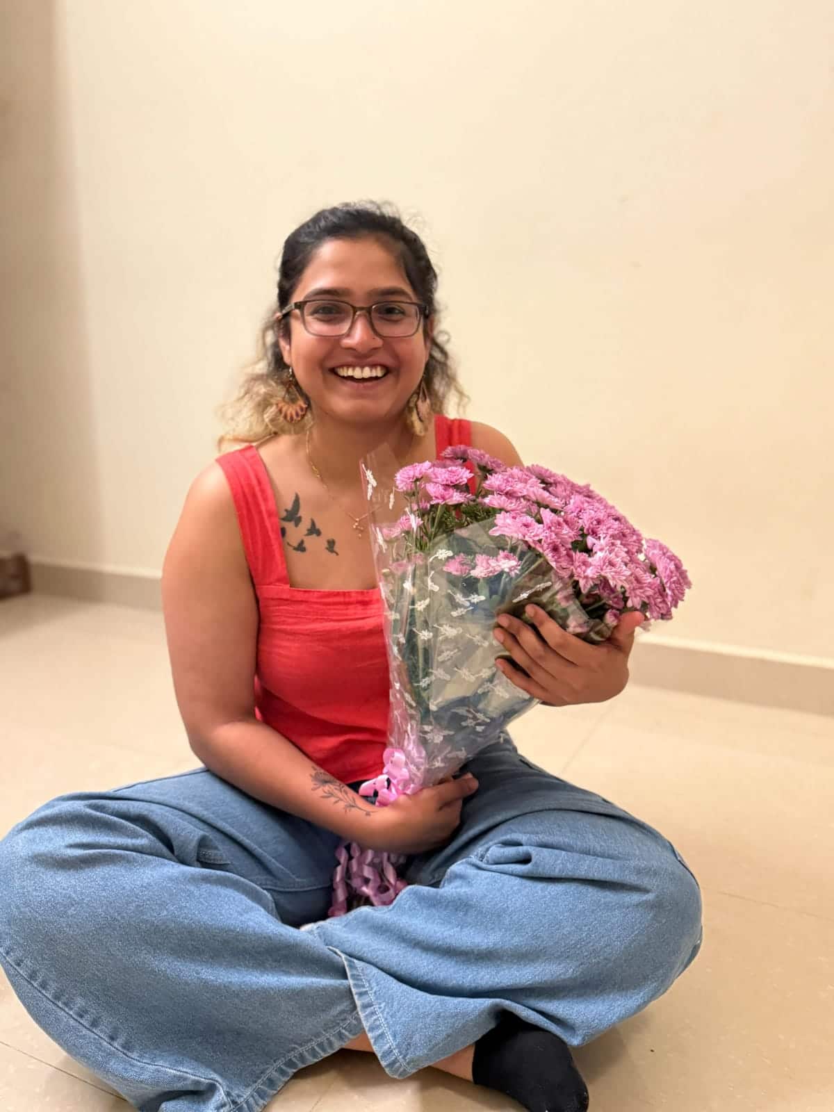 A smiling woman wearing blue jeans and a red sleeveless top sits cross-legged on the floor and smiles. She is holding a bunch of pink flowers.