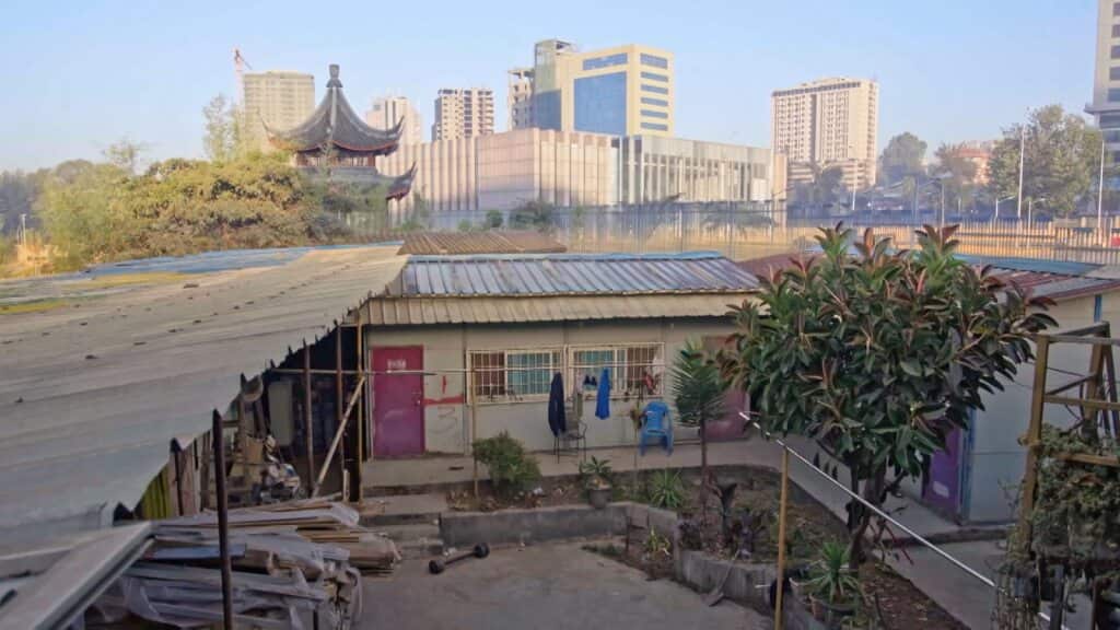 A house with a corrugated iron roof and backyard in the foreground, with tall office buildings and a temple behind it. 