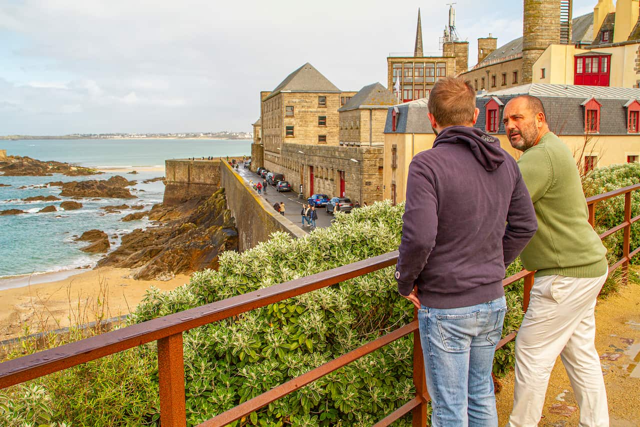 Two men lean on a fence, overlooking a sweepiing view of a walled city overlooking the sea. 