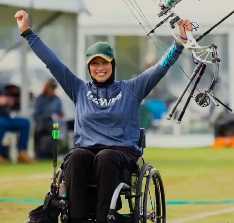 A woman raises her arms in celebration with a bow and arrows in her hands.