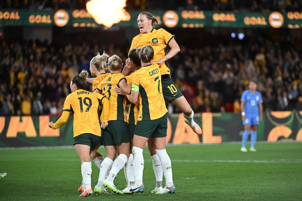 Women in yellow soccer shirts and green shorts celebrate in a huddle. One jumps up and pushes herself over the top.