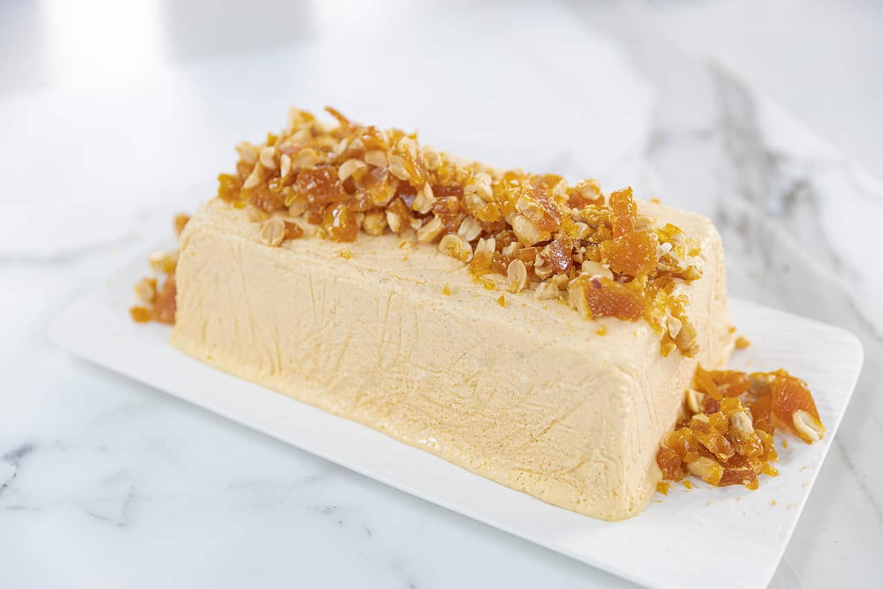 A long block of cremy coloured parfait sits on a white platter. A line of peanut brittle pieces sits along the top of the parfait. 