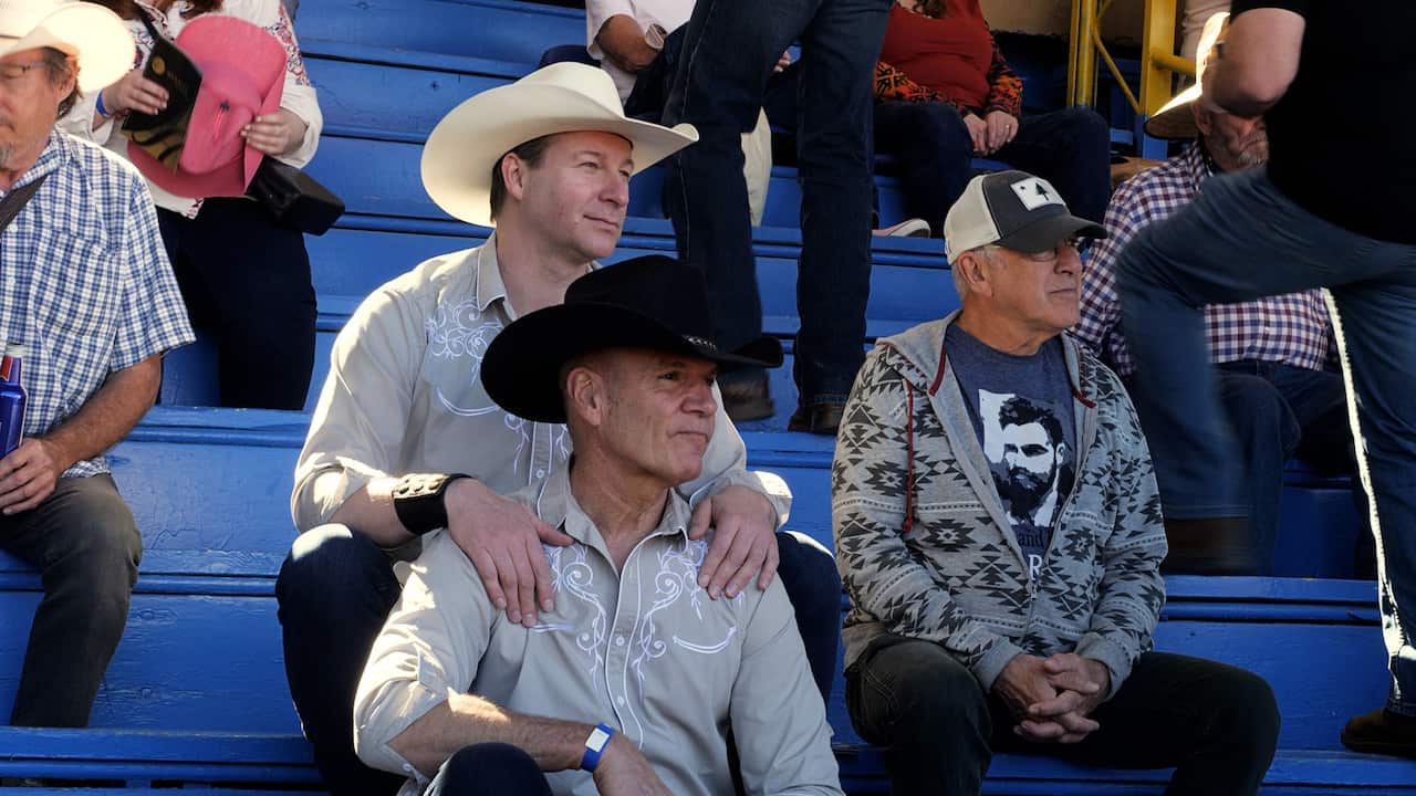 Two men in cowboy hats sit among spectators on a sports arena benches. One rests his hands on the other's shoulders