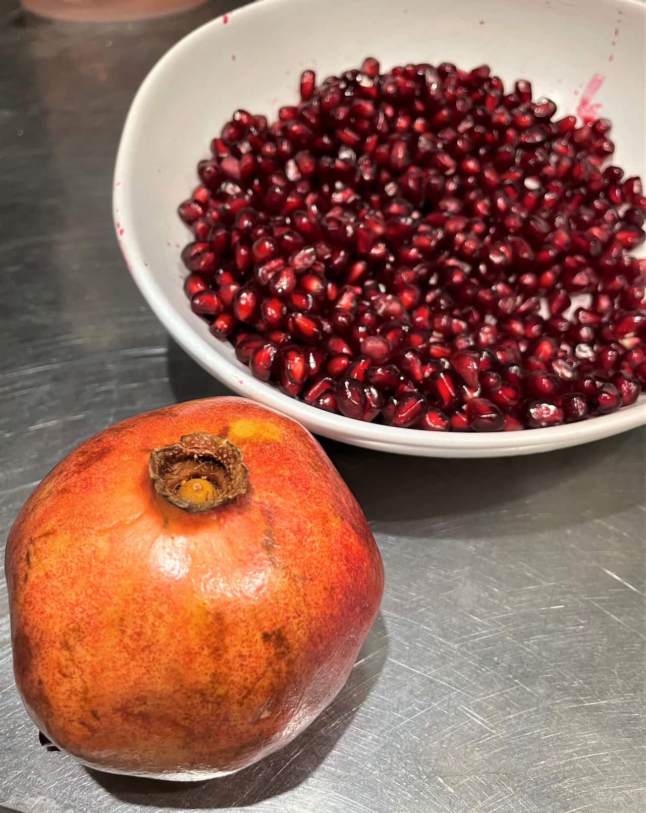 A whole fresh pomegranate, and a bowl of seeds on a bench.