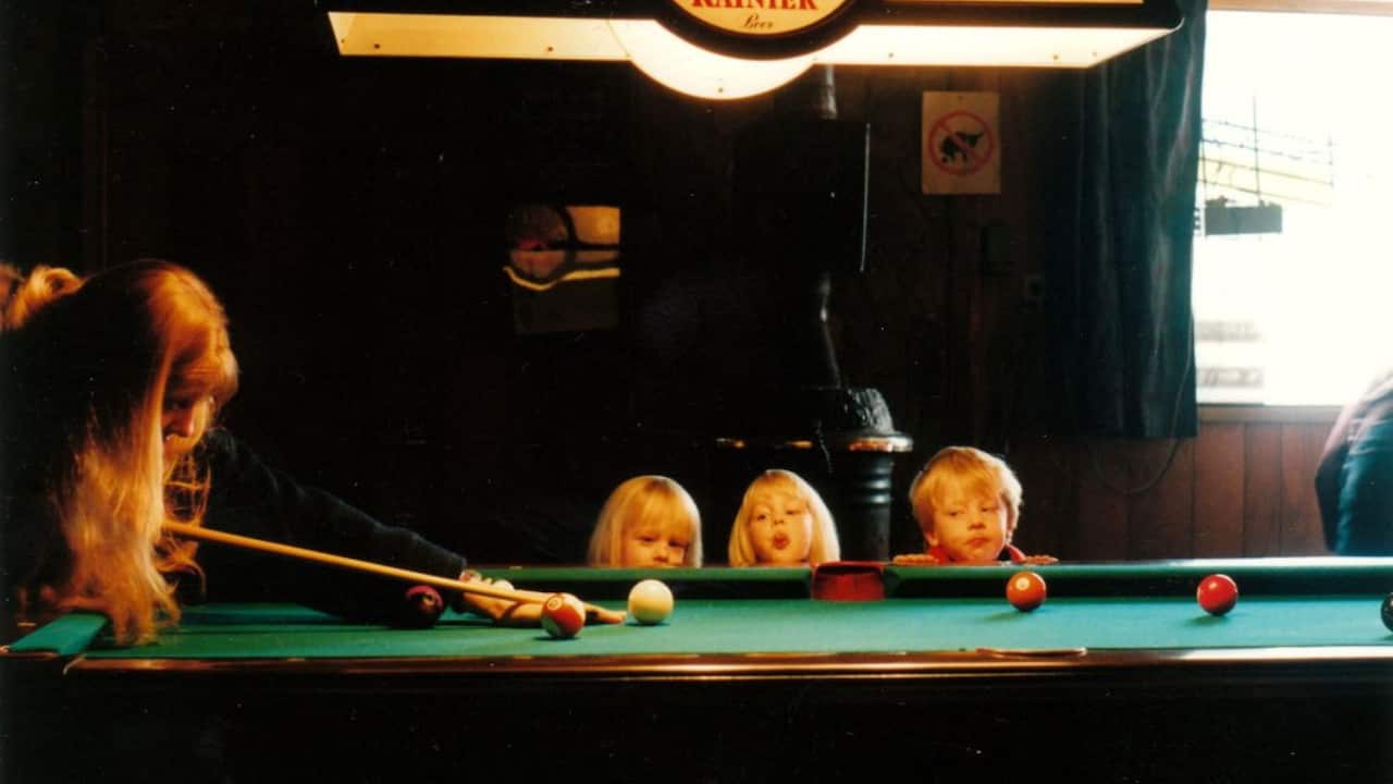 a woman with long blonde hair plays pool with three short young blonde children looking on
