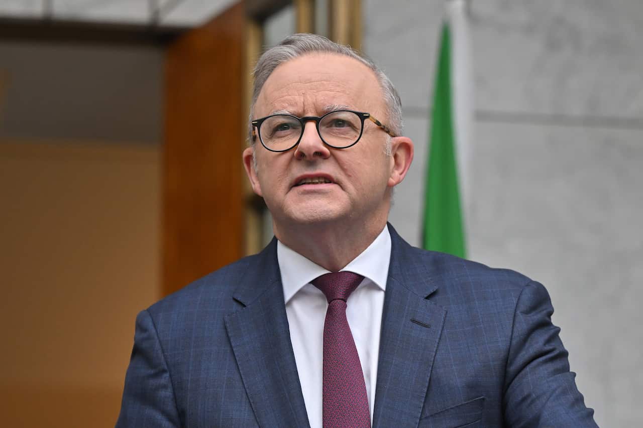 Anthony Albanese, wearing a blue suit, white shirt, and red tie stands outdoors with a flag in the background.