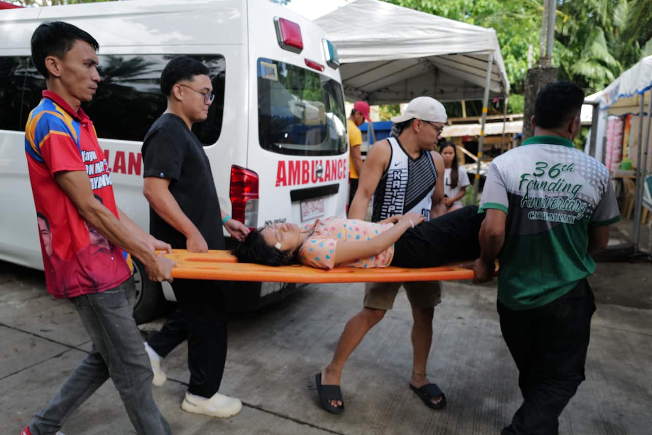 A woman on a stretcher is carried by four men, an ambulance is behind her