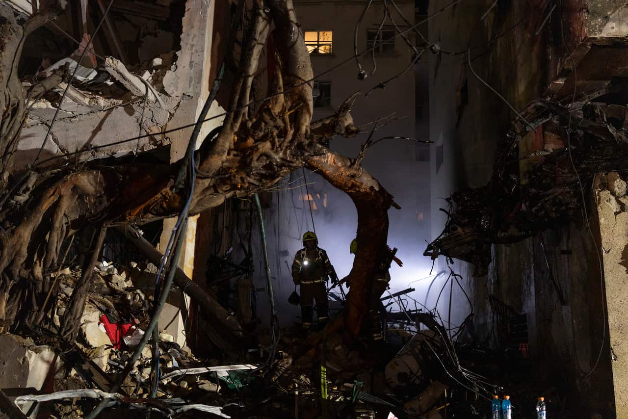 Firefighters with headlamps search through heavy rubble and twisted debris inside a damaged building at night.