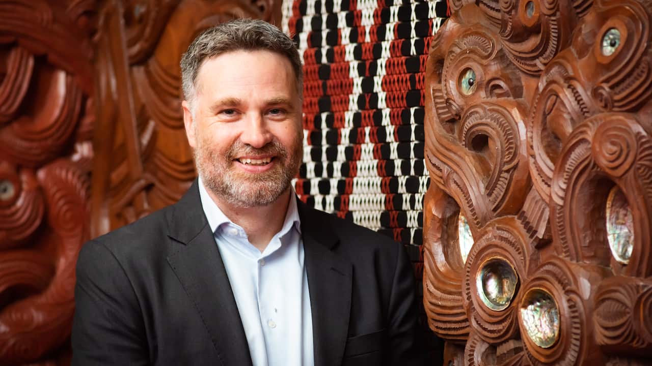 A middle-aged man wearing a black suit jacket sits in front of a carved wood surface. 
