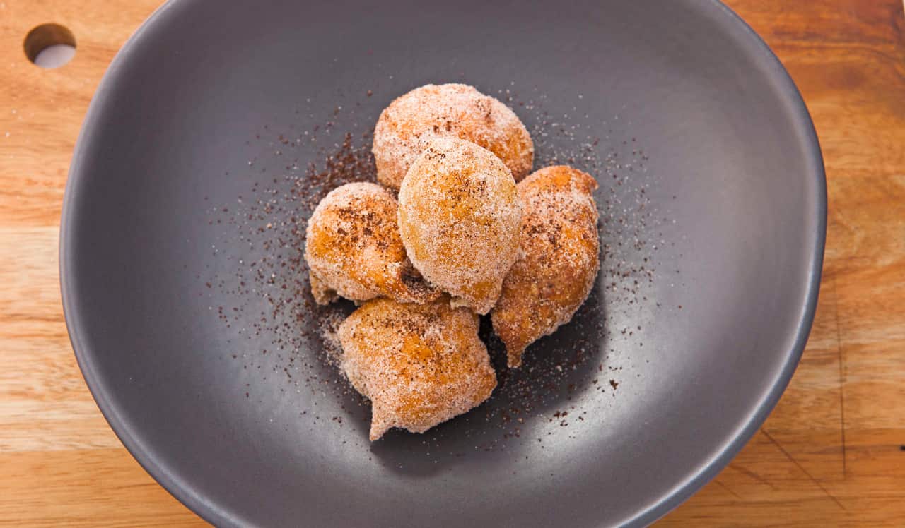 Five golden bown, sugar-dusted doughnut balls sit in a dark grey plate.
