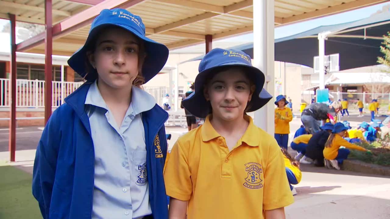 Two young girls wearing blue hats and school uniforms are standing in an open space outdoors.