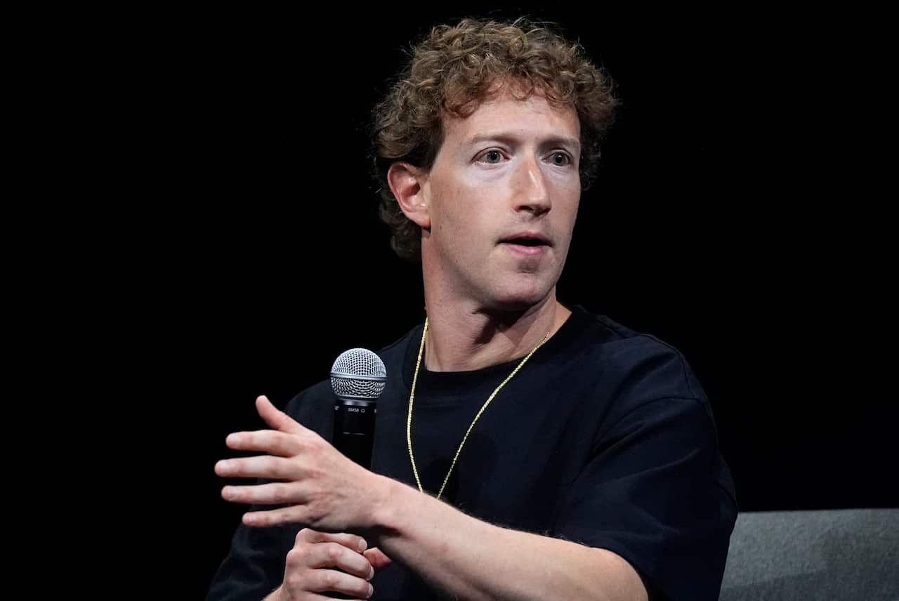 Mark Zuckerberg in a black shirt with a gold chain speaking into a microphone
