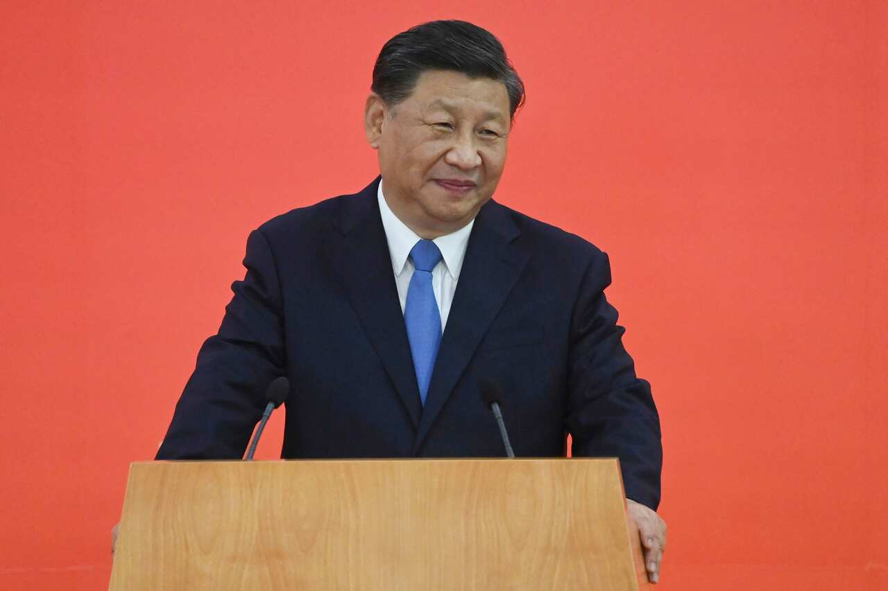 Xi Jinping stands at a podium in a suit and blue tie, with a small smile. 