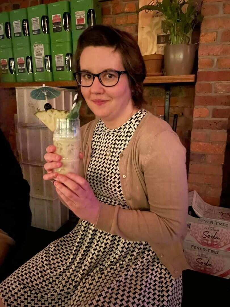 A woman wearing a dress and cardigan is sitting down and drinking a cocktail.