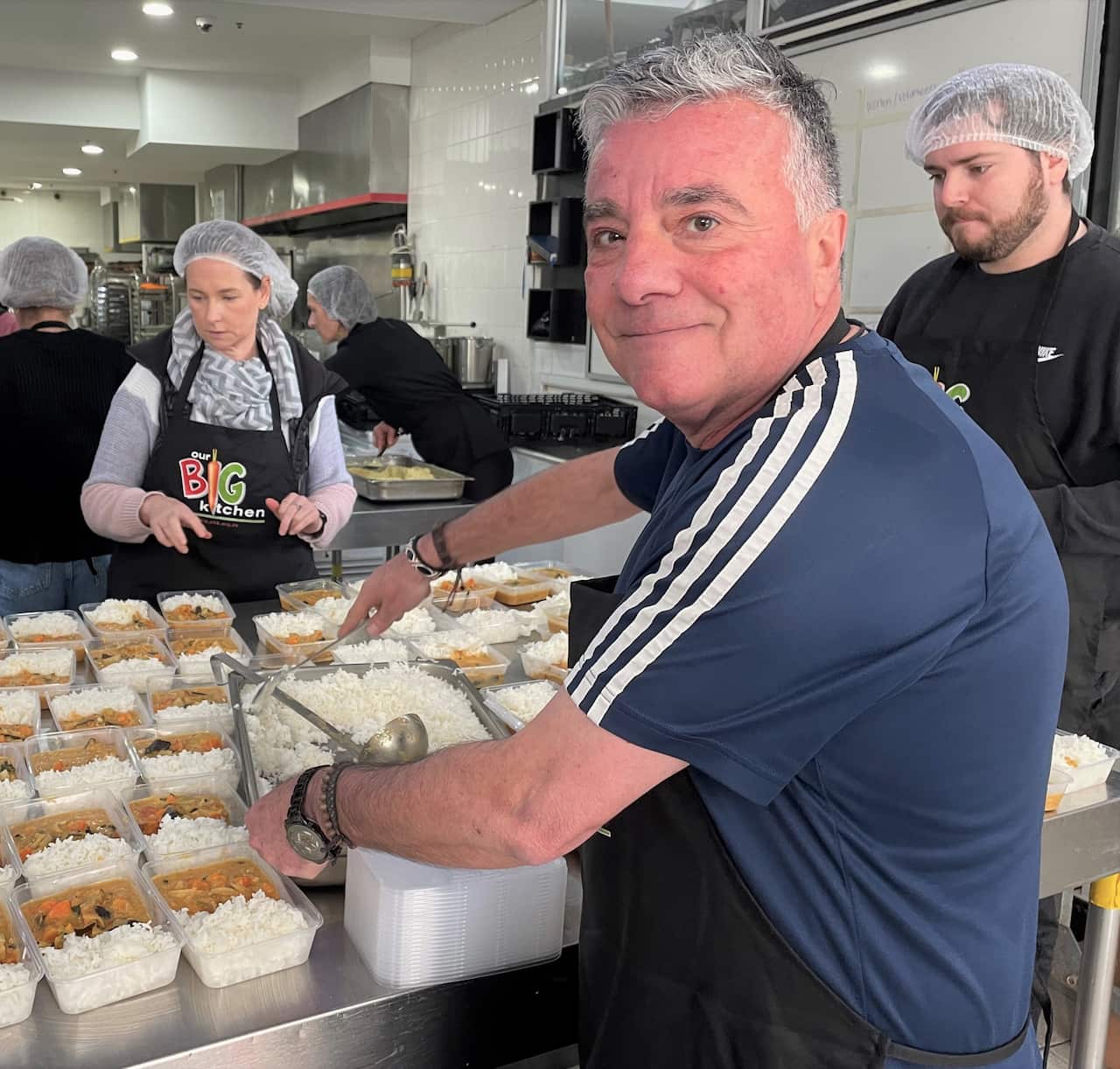 A man in a blue t-shirt faces camera while putting rice into meal boxes in a commercial kitchen.