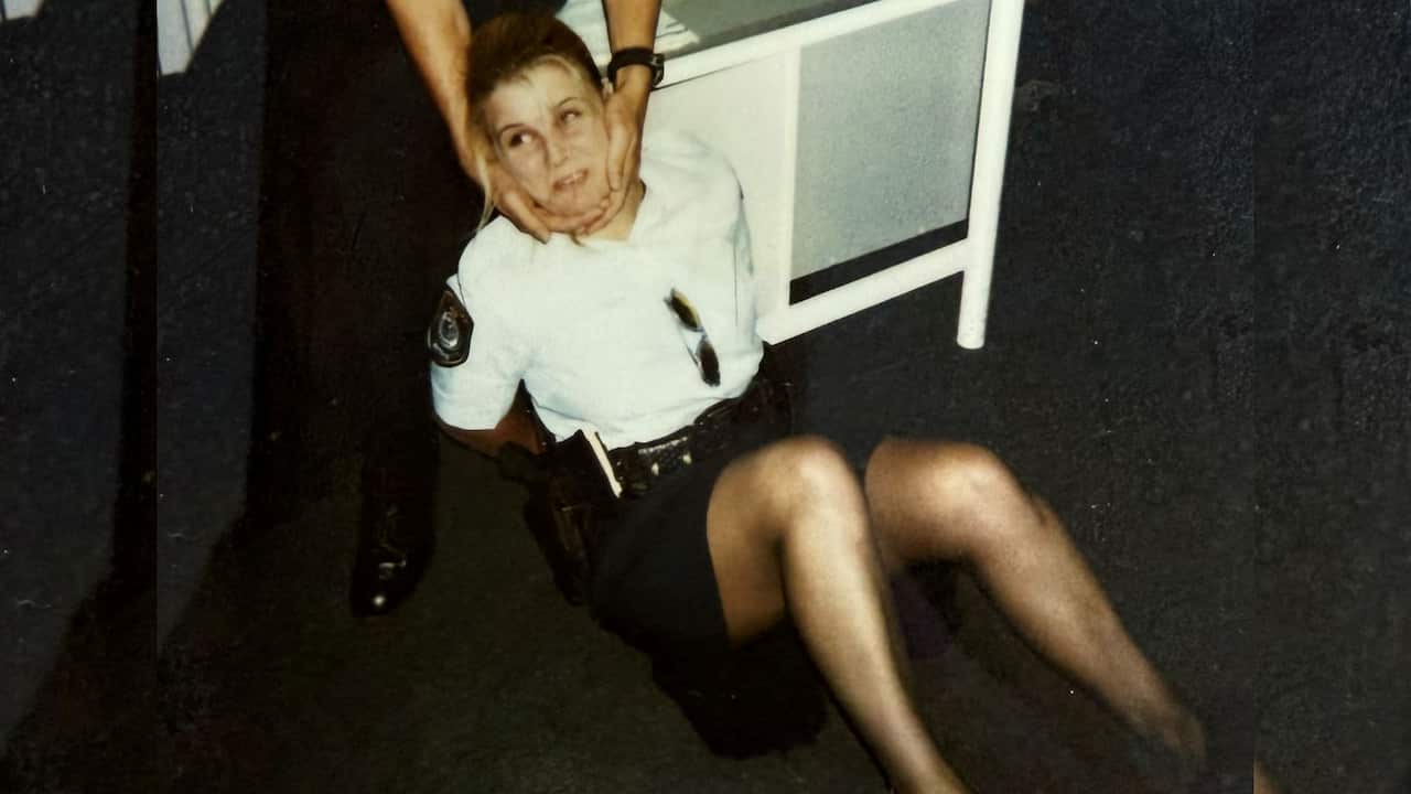 a young female police officer is sitting on the floor handcuffed to a desk with the arms of a male officer holding her face to face the camera